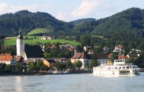 Stadtansicht mit Kirche und Schiff auf einem Fluss, umgeben von Hügeln., © Donau Schiffsstationen GmbH